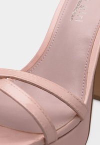 Prim-plan cu o sandală roz pal, cu vârf deschis, cu două curele și talpă cusută, arătând o parte din toc bloc.