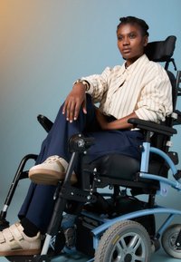 Fauteuil roulant électrique avec un cadre bleu et des sièges noirs. Personne portant une chemise claire à rayures et un pantalon marine, associé à des chaussures beiges.