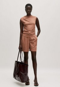 Brauner, ärmelloser Top mit einem verdrehten Design, kombiniert mit passenden Shorts. Halte eine dunkelburgunderfarbene Tragetasche. Einfacher, glatter Stoff.