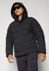 Giacca a vento nera con cappuccio, caratterizzata da un design trapuntato, tasche con zip e una texture liscia. Indossata sopra pantaloni scuri.