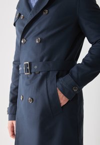 Gabardina de color azul marino con doble botonadura, cinturón en la cintura, grandes botones y bolsillos laterales. Tejido suave con un ajuste entallado.