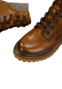 Brązowe skórzane botki z szarą teksturowaną tkaninową wykładziną przy kostce, okrągłymi sznurowadłami i grubą, wytrzymałą gumową podeszwą zapewniającą przyczepność.