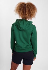 Sudadera con capucha verde hecha de tejido suave, con un diseño ajustado y puños acanalados. Llevada con pantalones cortos negros, mostrando la vista trasera.