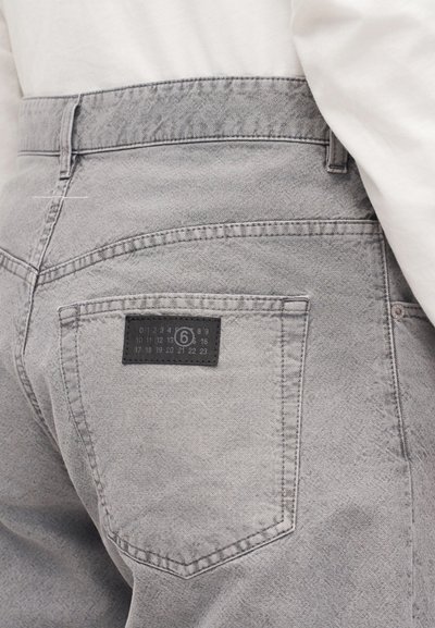 Γκρι τζιν παντελόνι από denim με τσέπη στη ράχη με ραφές και μαύρη ετικέτα που εμφανίζει αριθμούς και λογότυπο "6" σε κύκλο, φορεμένο με λευκό τοπ.