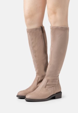 Bottes en suède beige jusqu'aux genoux avec une fermeture éclair sur le côté et un petit talon empilé, portées sur des jambes à la peau claire sur un fond blanc.