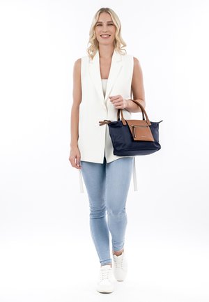 Sac en bandoulière bleu marine avec des accents en cuir marron. Tissu doux, forme rectangulaire, deux poignées et une poche zippée à l'avant. Tenue décontractée avec un jean clair.