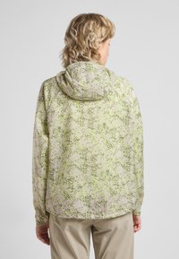 Veste légère à capuche avec un motif tacheté vert et beige. Elle présente une coupe décontractée et de longues manches avec des poignets élastiques.