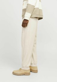 Beige sweatpants med elastiske manchetter, lavet af blødt stof. Båret med lysebrune snørresko, set fra siden. Simpelt design uden mønstre.