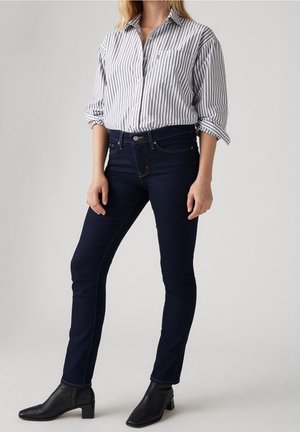 Femme portant un jean skinny foncé, des bottines noires et une chemise blanche à rayures verticales noires, debout devant un fond uni.