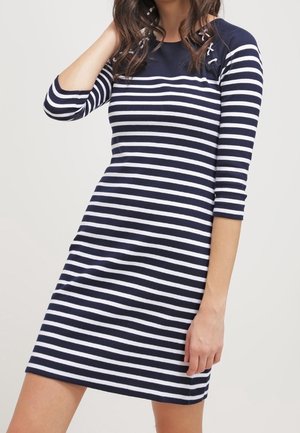 Kobieta w granatowo-białej sukience w poziome paski z rękawami do połowy przedramienia, dotykająca ręką włosów, na jasnym tle.