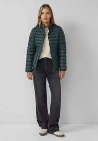 Doudoune vert foncé avec un design matelassé léger, col montant et fermeture éclair, associée à un jean gris à jambe large et des baskets blanches.