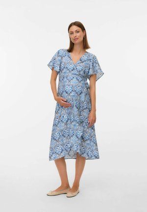 Donna incinta in piedi,che indossa un vestito avvolgente a righe blu e ballerine beige, mentre tiene delicatamente il suo pancione contro uno sfondo bianco.