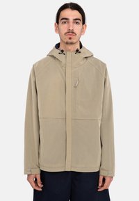 Chaqueta beige con un patrón y capucha, con cremallera frontal, cordones ajustables y bolsillos, hecha de un material ligero.