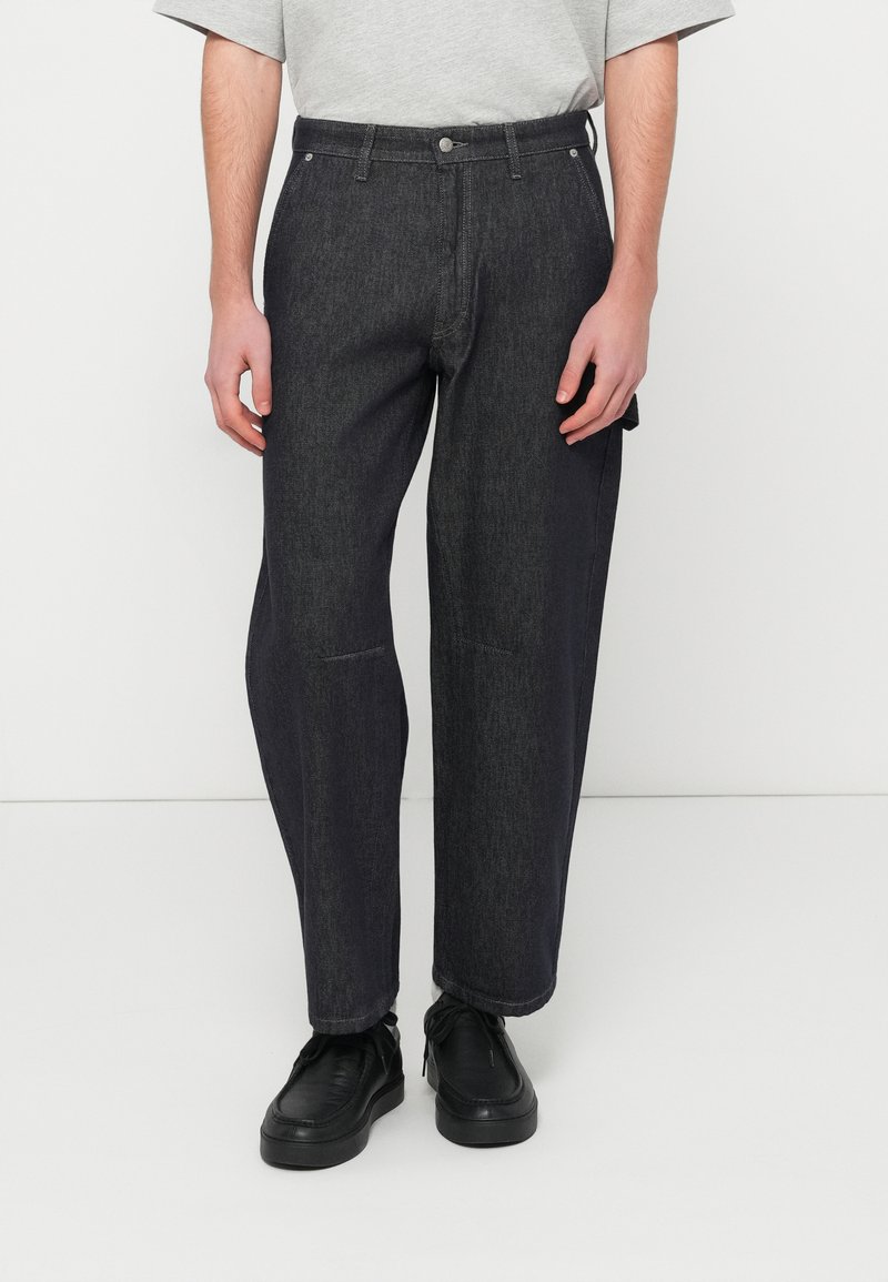 Temno modre kavbojke z ohlapnim krojem in ravnimi hlačnicami, opremljene z gumbom in zadrgo, stranskimi žepi ter minimalnimi šivi.