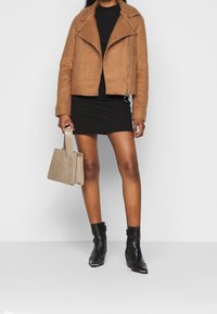 Kvinna iklädd en brun jacka, svart klänning, svarta ankelskor, som håller en beige texturerad väska och står mot en enfärgad bakgrund.