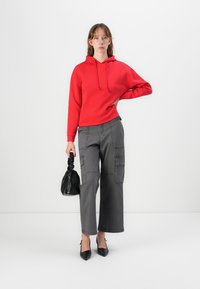 Sarkans kapuci krekls ar aukliņu, apvienots ar pelēkiem platiem biksēm ar kabatām. Tur rokās mazu melnu somiņu, uz kājām melnas kurpes ar papēžiem.