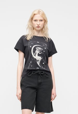 Blonde Frau trägt ein schwarzes T-Shirt mit einer weißen Meerjungfrau, die auf einem Halbmond sitzt, und schwarze Shorts vor einem einfarbigen Hintergrund.