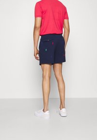 Shorts de color navy con logotipos bordados coloridos, dos bolsillos traseros y cierre de botón, combinados con una camiseta de manga corta roja y zapatillas blancas.