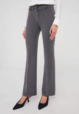 Donna in pantaloni grigi a gamba dritta, camicia bianca a maniche lunghe e décolleté nere a punta, in piedi su uno sfondo neutro.