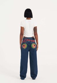 Camiseta blanca corta combinada con unos vaqueros de denim azules de corte holgado que presentan coloridos diseños bordados en los bolsillos y letras rojas de "Ed Hardy".