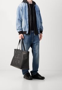 Denim jacket over black hoodie, paired with blue jeans and black sneakers. Black textured tote bag with embossed "HUGO" logo.