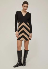 Abito nero a maniche lunghe con scollatura a V, caratterizzato da una parte inferiore beige decorata con audaci strisce a zigzag nere. Stivali neri fino al ginocchio.