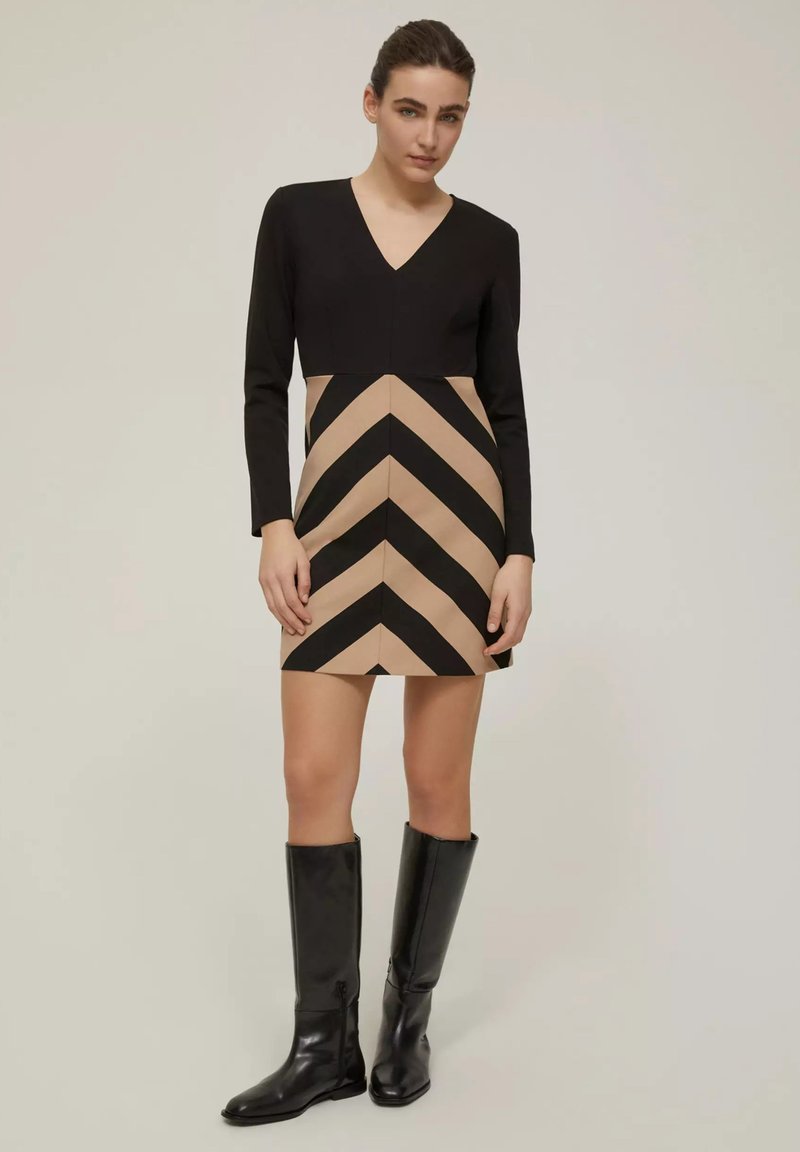 Abito nero a maniche lunghe con scollatura a V, caratterizzato da una parte inferiore beige decorata con audaci strisce a zigzag nere. Stivali neri fino al ginocchio.
