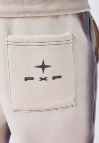 Tasca di pantaloni beige con design a stella nera e testo "PXP" stampato sul tessuto, cucito con filo nero visibile.