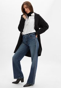 Hoch geschnittene, dunkelblaue Jeans aus Denim mit weitem Bein, verziert mit silbernen Nieten entlang der Seite, kombiniert mit einem weißen Hemd und einem schwarzen Mantel.
