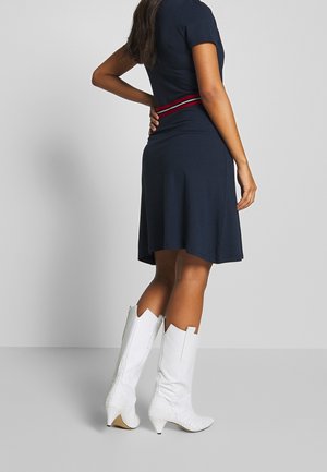 Femme portant une robe bleu marine avec une ceinture rayée rouge et blanche, et des bottes blanches à talons mi-mollet, debout avec une main posée sur le dos.