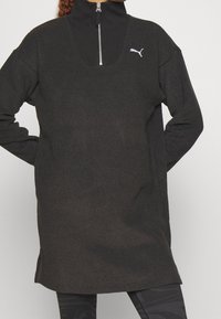 Persoon in een zwarte fleece pullover met lange mouwen en kwart rits, met het Puma-logo, gecombineerd met zwart gemusterde legging.