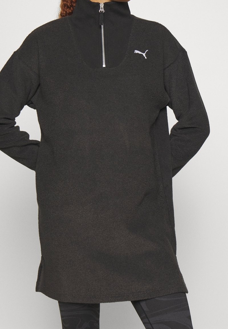 Persoon in een zwarte fleece pullover met lange mouwen en kwart rits, met het Puma-logo, gecombineerd met zwart gemusterde legging.