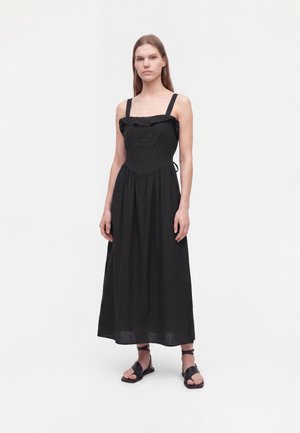 Femme debout, portant une longue robe noire sans manches avec un col à volants et des sandales plates noires à brides chevilles, sur fond blanc.