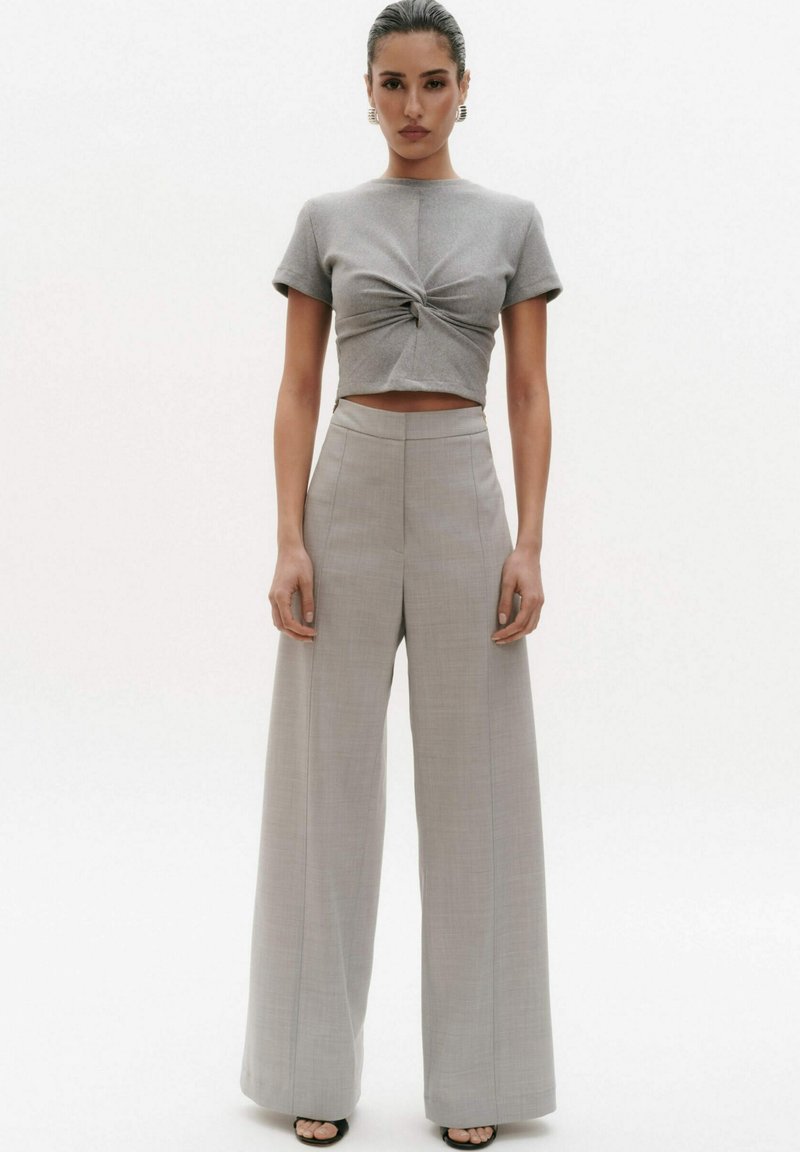 Szary crop top z wiązanym przodem, zestawiony z jasnoszarymi, szerokimi spodniami w wysokim stanie. Gładki materiał i dopasowany krój. Czarne sandały na obcasie z odkrytymi palcami.