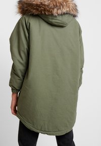 Olive green parka with a faux fur-trimmed hood, long sleeves, elastic cuffs, and a curved hem. Smooth fabric with a casual design.