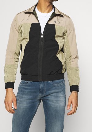 Mann trägt eine farblich unterteilte beige, olivegrüne und schwarze Jacke mit Reißverschluss, schwarzen Bündchen und blauen Jeans, steht vor einem einfarbigen Hintergrund.