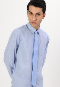 Camisa de vestir a rayas azules con mangas largas y cuello abotonado, combinada con una corbata azul claro. Presenta un bolsillo en el pecho con una etiqueta.