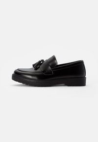 Mocassins en cuir noir avec une semelle plate, ornés d'un gland décoratif sur le devant et de détails cousus le long des bords.