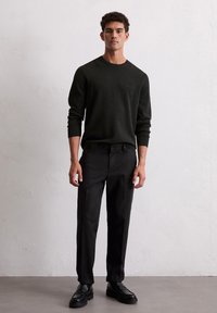 Homme se tenant contre un mur blanc, portant un pull noir à manches longues, un pantalon noir et des chaussures en cuir noires, faisant face à l'avant les mains sur les côtés.