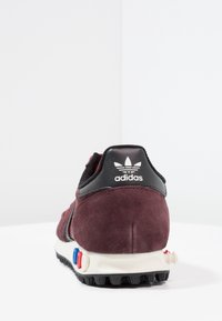 Bordeaux mocka sneaker med svarta läderdetaljer, med en vit mellansula och färgglad knappdetalj på hälen. Klassisk tre ränder design.