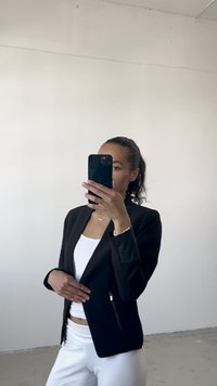 Fekete blézer kontrasztos bőrhatású ujjal, slim fit fazon, egyetlen cipzáras zsebbel, fehér felsőn viselve, fehér nadrággal kombinálva.