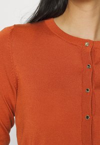 Femme portant un cardigan en tricot orange rouille avec des boutons ronds en or, gros plan sur l'épaule et l'encolure, cheveux foncés visibles.