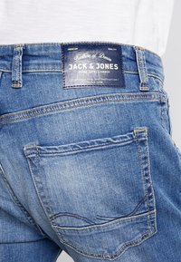 Jeans de mezclilla azul con una etiqueta de cuero con la marca "Jack & Jones", detalles en el bolsillo trasero y una textura desgastada.