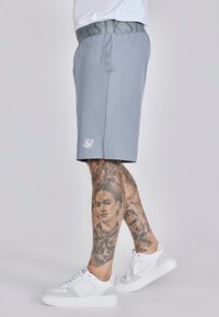 Gray athletic shorts with a wide elastic waistband featuring a logo. The fabric appears smooth and lightweight. Worn with white sneakers.