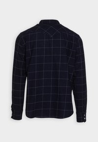 Marinblå rutiga skjorta med vitt rutnät, långa ärmar, knäppningar vid ärmsluten och en klassisk krage. Har en axeldesign på baksidan.