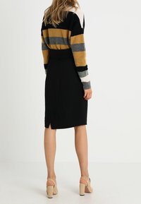 Suéter de punto a rayas en negro, mostaza, gris y blanco; falda negra ajustada con una abertura en la parte posterior; complementado con zapatos de tacón alto beige.