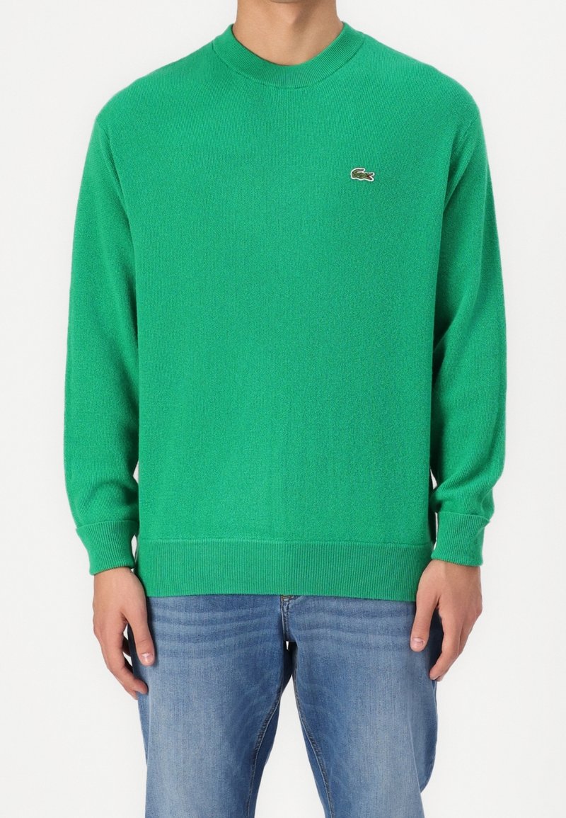 Homme portant un sweat-shirt vert vif col rond avec un petit logo crocodile brodé sur la poitrine gauche et un jean bleu.