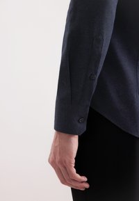 Ciemnoniebieska, fakturowana koszula z długim rękawem, z zapięciem na guziki przy mankietach oraz subtelnymi detalami wzoru, prezentowana na dłoni na jasnym tle.