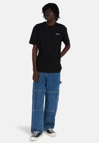 Camiseta de algodón negro con logotipo, combinada con pantalones cargo de denim azul anchos con diseño de patchwork y bolsillos laterales. Zapatillas negras.