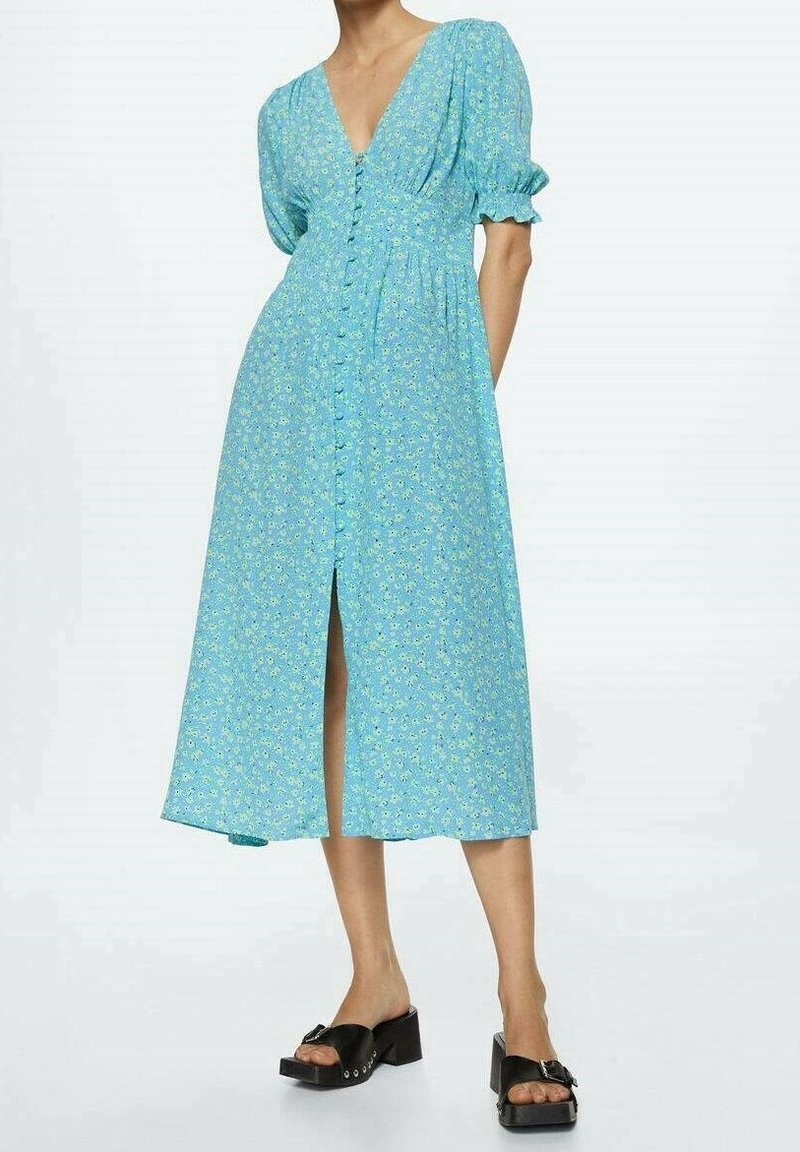 Femme portant une robe midi bleue à motifs floraux avec des boutons sur le devant, des manches bouffantes, des poches latérales et des sandales noires à talons carrés.